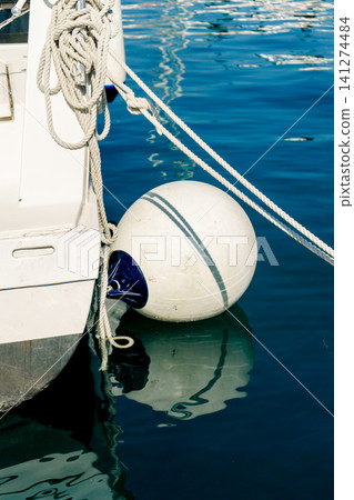 White yacht fender protects a moored boat as calm marina water reflects ropes and hull, capturing nautical safety and harbor life White yacht fender protects a moored boat as calm marina water reflects ropes and hull, capturing nautical safety and harbor life 141274484