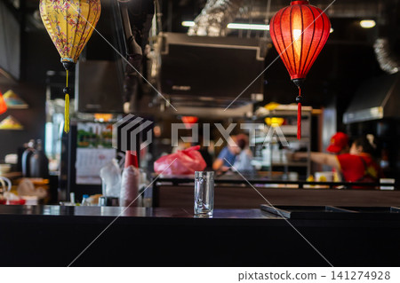Empty table with glass tumbler and lit lanterns in bustling Asian restaurant interior at night Empty table with glass tumbler and lit lanterns in bustling Asian restaurant interior at night 141274928