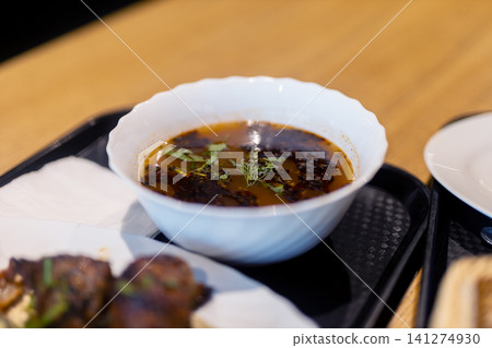 Dark dipping sauce in white scalloped bowl with herb garnish on black tray beside grilled meat skewers over rustic wooden table, with sauce, dipping, soy, bowl detail Dark dipping sauce in white scalloped bowl with herb garnish on black tray beside grilled meat skewers over rustic wooden table, with sauce, dipping, soy, bowl detail 141274930