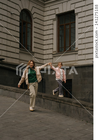 A mother and daughter walk together arm in arm on a street near a building in the city during the day. A mother and daughter walk together arm in arm on a street near a building in the city during the day. 141275188