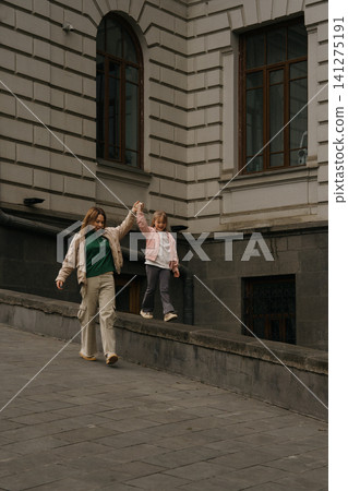 Mom walks hand in hand with daughter on city street near building in daytime Mom walks hand in hand with daughter on city street near building in daytime 141275191