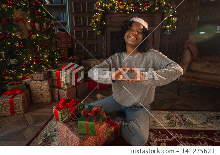 Merry Christmas. African American woman smiling making heart shape with hands near Christmas tree. Happy girl in Santa hat in living room with fireplace Christmas gift boxes. Christmas eve at home Merry Christmas. African American woman smiling making heart shape with hands near Christmas tree. Happy girl in Santa hat in living room with fireplace Christmas gift boxes. Christmas eve at home 141275621