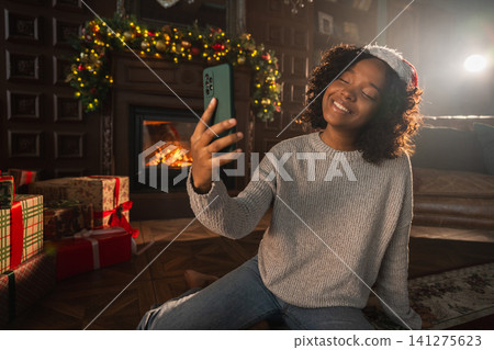 Merry Christmas. African woman in Santa hat having video chat in smartphone. Blogger in festive mood recording video in social network. Girl having video call with friends family celebrating Christmas Merry Christmas. African woman in Santa hat having video chat in smartphone. Blogger in festive mood recording video in social network. Girl having video call with friends family celebrating Christmas 141275623
