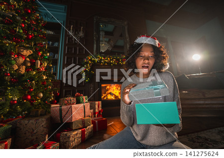 Merry Christmas. African American woman unpacking open up gift box near Christmas tree. Girl in living room with Christmas tree and fireplace opening gift box with surprise face. Christmas eve at home Merry Christmas. African American woman unpacking open up gift box near Christmas tree. Girl in living room with Christmas tree and fireplace opening gift box with surprise face. Christmas eve at home 141275624