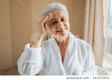 Mature adult old woman doing daily beauty routine removing makeup with cotton pad. Senior woman with lotion sponge washing face gets facial treatment. Skin care aging beauty skincare cleansing Mature adult old woman doing daily beauty routine removing makeup with cotton pad. Senior woman with lotion sponge washing face gets facial treatment. Skin care aging beauty skincare cleansing 141275643