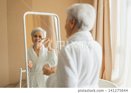 Mature adult old woman with smooth soft perfect skin applying rouge with brush on cheek looking at mirror doing daily beauty make up routine. Senior woman putting blush on her face. Anti age makeup Mature adult old woman with smooth soft perfect skin applying rouge with brush on cheek looking at mirror doing daily beauty make up routine. Senior woman putting blush on her face. Anti age makeup 141275644