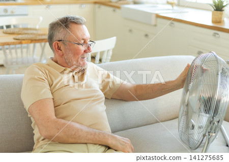 Senior man refreshing himself near fan during summer heat wave hot day. Elderly man suffering from summer heat trying to survive with electric fan. Man enjoying refreshing air blowing from ventilator Senior man refreshing himself near fan during summer heat wave hot day. Elderly man suffering from summer heat trying to survive with electric fan. Man enjoying refreshing air blowing from ventilator 141275685