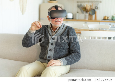 Excited mature senior man wearing using virtual reality metaverse VR glasses headset at home. Grandfather touching air during VR experience on virtual reality helmet. Simulation hi-tech videogame 141275696