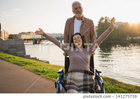 Senior woman in wheelchair walking with caregiver old man on road. Family couple man husband supporting taking care of paralyzed woman wife in chair for people with disability outdoor. Rehabilitation Senior woman in wheelchair walking with caregiver old man on road. Family couple man husband supporting taking care of paralyzed woman wife in chair for people with disability outdoor. Rehabilitation 141275739