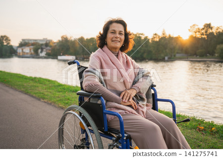 Happy senior woman in wheelchair smiling enjoying freedom walking on road in city park outdoor. Old cheerful positive woman in chair for people with disability. Disability disease rehabilitation Happy senior woman in wheelchair smiling enjoying freedom walking on road in city park outdoor. Old cheerful positive woman in chair for people with disability. Disability disease rehabilitation 141275741