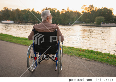 Sad senior man sitting in wheelchair walking on road in city park outdoor, back view. Old paralyzed man in chair for people with disability feel depressed. Lonely elderly sick man sits on wheelchair Sad senior man sitting in wheelchair walking on road in city park outdoor, back view. Old paralyzed man in chair for people with disability feel depressed. Lonely elderly sick man sits on wheelchair 141275743