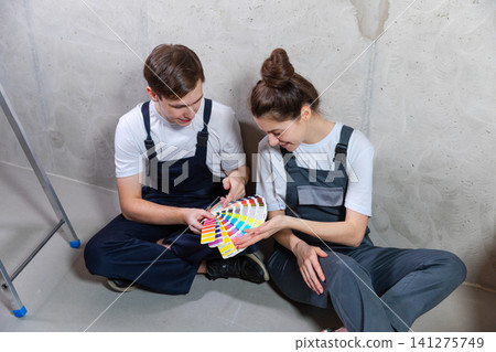 House decoration DIY renovation concept. Happy couple sitting on floor in empty room of new home planning design. Husband and wife holding swatch palette choosing colors for painting wall in new house 141275749
