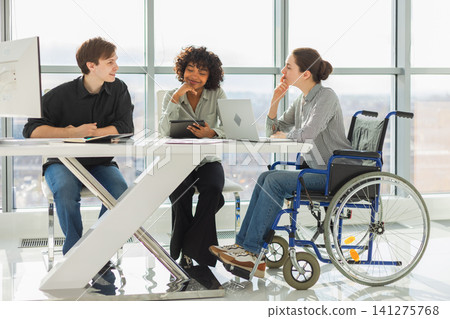 Teamwork brainstorm. Diverse multiracial colleagues woman in chair for people with disability working together. Group of people sitting in creative office discussing business. Young professional team Teamwork brainstorm. Diverse multiracial colleagues woman in chair for people with disability working together. Group of people sitting in creative office discussing business. Young professional team 141275768