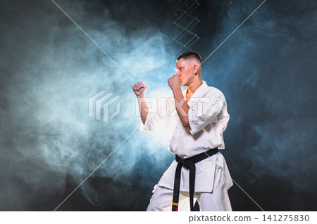 Strong man fighter in white kimono for karate martial arts with black belt preparing for fight training in black background. Man training moves punches kicks practicing shadow fight ready for sparring Strong man fighter in white kimono for karate martial arts with black belt preparing for fight training in black background. Man training moves punches kicks practicing shadow fight ready for sparring 141275830