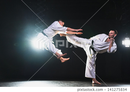 Martial arts masters on dark background. Strong man fighters in white kimono for karate martial arts fighting training in black gym. Karate fighters training punches kicks practicing fight sparring Martial arts masters on dark background. Strong man fighters in white kimono for karate martial arts fighting training in black gym. Karate fighters training punches kicks practicing fight sparring 141275834
