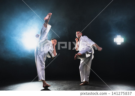 Martial arts masters on dark background. Strong man fighters in white kimono for karate martial arts fighting training in black gym. Karate fighters training punches kicks practicing fight sparring Martial arts masters on dark background. Strong man fighters in white kimono for karate martial arts fighting training in black gym. Karate fighters training punches kicks practicing fight sparring 141275837