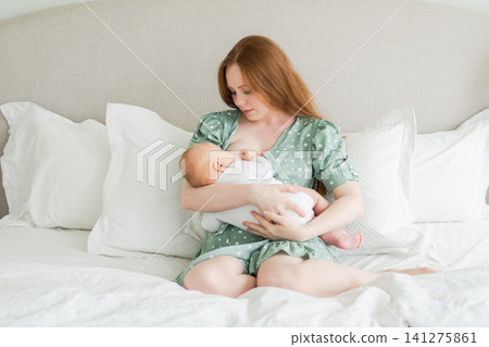 Happy family at home. Mother breastfeeding baby for health nutrition wellness. Mom nursing feeding her newborn child milk at home. Woman infant girl in white bedroom Mother hugs baby with love care Happy family at home. Mother breastfeeding baby for health nutrition wellness. Mom nursing feeding her newborn child milk at home. Woman infant girl in white bedroom Mother hugs baby with love care 141275861