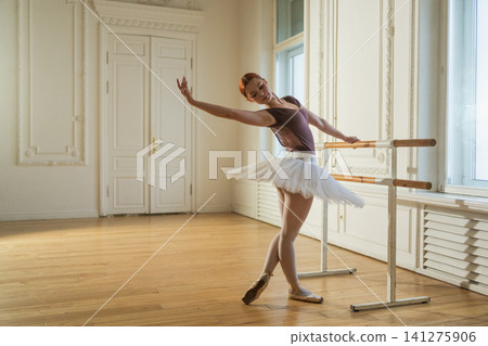 Young ballerina in tutu skirt stretching practicing ballet positions dancing near ballet barre in dance class. Young classical dancer woman in dance studio. Ballet class training Ballet school concept Young ballerina in tutu skirt stretching practicing ballet positions dancing near ballet barre in dance class. Young classical dancer woman in dance studio. Ballet class training Ballet school concept 141275906