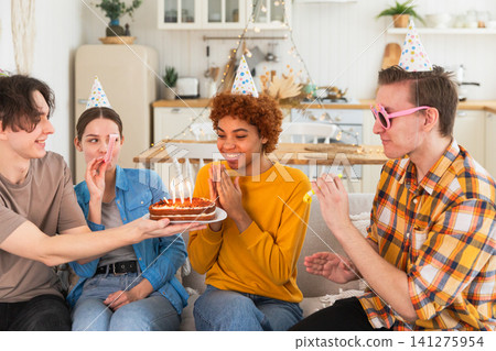 Make a wish. Woman wearing party cap blowing out burning candles on birthday cake. Happy Birthday party. Group of friends wishes girl happy birthday. People celebrating birthday with party at home Make a wish. Woman wearing party cap blowing out burning candles on birthday cake. Happy Birthday party. Group of friends wishes girl happy birthday. People celebrating birthday with party at home 141275954