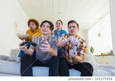 Home party. Cheerful group of friends playing video games at home. Happy diverse group buddies having fun together indoor. Friendship leisure entertainment concept. Young best friends enjoying weekend Home party. Cheerful group of friends playing video games at home. Happy diverse group buddies having fun together indoor. Friendship leisure entertainment concept. Young best friends enjoying weekend 141275958