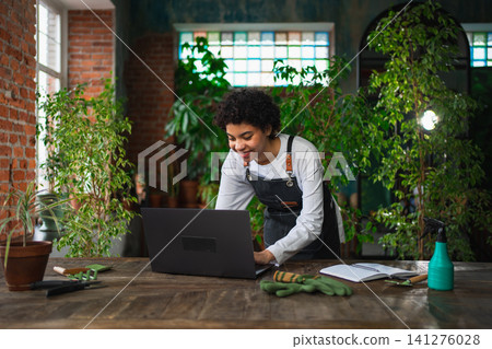 African woman florist gardener working in botanical store using laptop order plants communicating on Internet with customer. Female small business owner working at flower shop. Running of own business 141276028