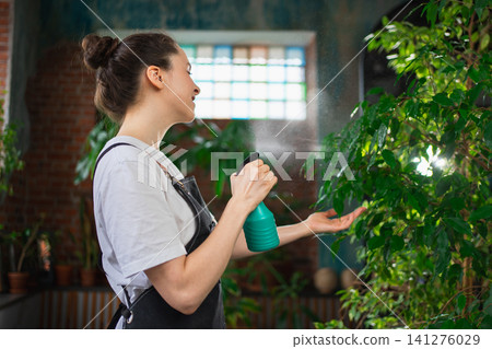 Running of own business. Young woman florist with spray bottle watering fertilizing plants in botanical store. Small business owner working at flower shop smiling surrounded by plants Small business 141276029