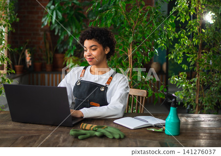 African woman florist gardener working in botanical store using laptop order plants communicating on Internet with customer. Female small business owner working at flower shop. Running of own business African woman florist gardener working in botanical store using laptop order plants communicating on Internet with customer. Female small business owner working at flower shop. Running of own business 141276037