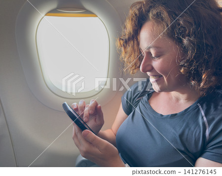 Passenger woman is flying in plane. Girl using phone sitting by airplane window. Traveling female with smartphone inside plane enjoying flight. Woman using wifi connection on board. Traveling girl Passenger woman is flying in plane. Girl using phone sitting by airplane window. Traveling female with smartphone inside plane enjoying flight. Woman using wifi connection on board. Traveling girl 141276145