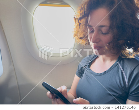 Passenger woman is flying in plane. Girl using phone sitting by airplane window. Traveling female with smartphone inside plane enjoying flight. Woman using wifi connection on board. Traveling girl 141276146