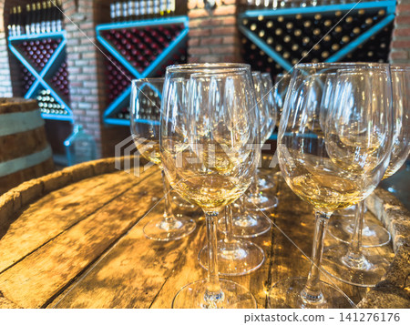 Many glasses of white wine on wooden barrel. Wine tasting event. Cellar with alcohol barrels. French wooden barrels. Wine shop bar winery or wine tasting concept Many glasses of white wine on wooden barrel. Wine tasting event. Cellar with alcohol barrels. French wooden barrels. Wine shop bar winery or wine tasting concept 141276176