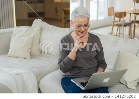 Confident stylish happy middle aged senior woman using laptop at home. Stylish older mature 60s grandmother sitting at couch looking at computer screen typing chatting reading writing email 141276208