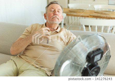 Senior man refreshing himself near fan during summer heat wave hot day. Elderly man suffering from summer heat trying to survive with electric fan. Man enjoying refreshing air blowing from ventilator Senior man refreshing himself near fan during summer heat wave hot day. Elderly man suffering from summer heat trying to survive with electric fan. Man enjoying refreshing air blowing from ventilator 141276243