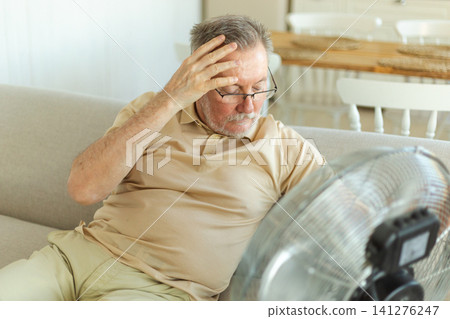 Senior man refreshing himself near fan during summer heat wave hot day. Elderly man suffering from summer heat trying to survive with electric fan. Man enjoying refreshing air blowing from ventilator 141276247