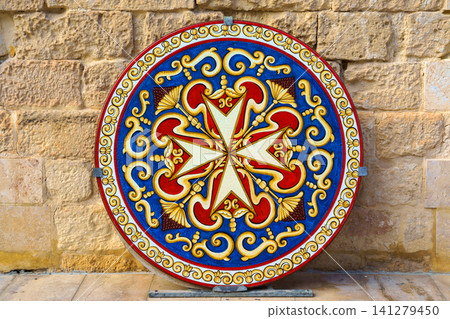 Valletta, Malta - Nov 7 2018, A close up view of a round shield with a colourful image of the Maltese Cross leaning against a brick wall, without people, at daytime, Valletta, Malta 141279450