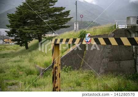 Amidst the rain, the landscape of the earthquake's relics—a height restriction sign warped and a rusted gate damaged by the tsunami during the Great East Japan Earthquake—presents a scene of disaster. Amidst the rain, the landscape of the earthquake's relics—a height restriction sign warped and a rusted gate damaged by the tsunami during the Great East Japan Earthquake—presents a scene of disaster. 141280570