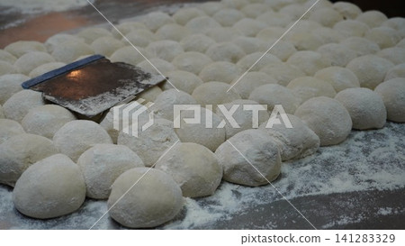 Many round balls of dough are floured, arranged on a surface, grouped together with a metal scraper laying on them. Many round balls of dough are floured, arranged on a surface, grouped together with a metal scraper laying on them. 141283329