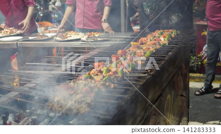 Skewered meat and vegetables grill over coals, smoke rising, people bustling in the background. Skewered meat and vegetables grill over coals, smoke rising, people bustling in the background. 141283333