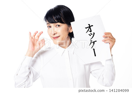 A middle-aged woman in a white shirt smiles at the camera, holding a piece of paper that says "OK". A middle-aged woman in a white shirt smiles at the camera, holding a piece of paper that says "OK". 141284099