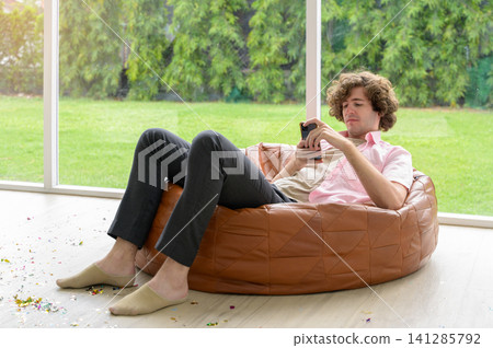 Relaxed young man using smartphone while sitting on bean bag chair in living room with large window and green garden view background Relaxed young man using smartphone while sitting on bean bag chair in living room with large window and green garden view background 141285792