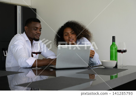 Young african couple using laptop together at home with wine glasses in modern kitchen Young african couple using laptop together at home with wine glasses in modern kitchen 141285793