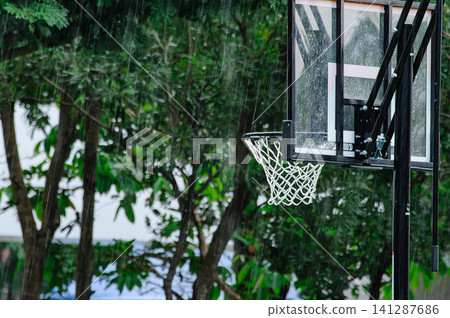 raining in the outdoor basketball field with a basketball hoop with many trees in background 141287686