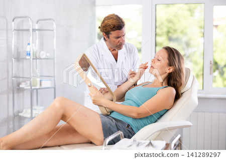 Woman looking at mirror while cosmetologist showing results of facial procedure Woman looking at mirror while cosmetologist showing results of facial procedure 141289297