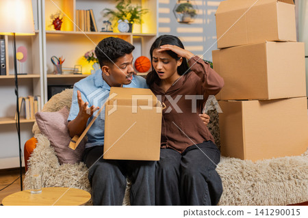 Indian couple man and woman sitting on home sofa opening delivery box and reacting to damaged order 141290015