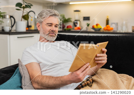 Middle-aged man reads book under plaid smiles enjoys free time comfort relax leisure chill rest Middle-aged man reads book under plaid smiles enjoys free time comfort relax leisure chill rest 141290020