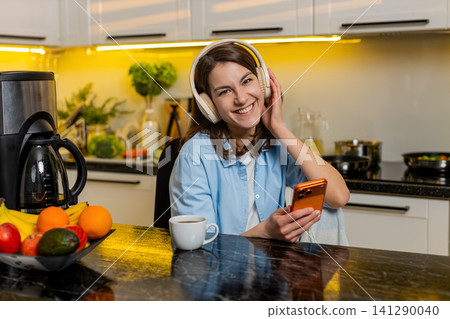 Young woman relaxing in home kitchen with headphones enjoying music while checking smartphone screen 141290040