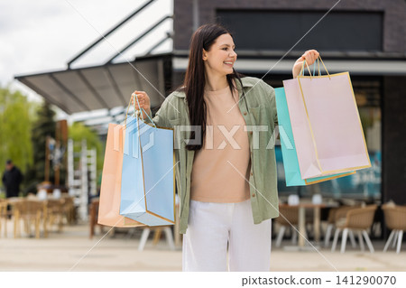 Young woman carrying shopping bags walking in city street smiling happily after successful shopping Young woman carrying shopping bags walking in city street smiling happily after successful shopping 141290070