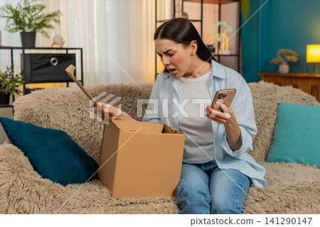 Young woman sitting on sofa unpacking delivery finding wrong item disappointed shaking head sadly Young woman sitting on sofa unpacking delivery finding wrong item disappointed shaking head sadly 141290147