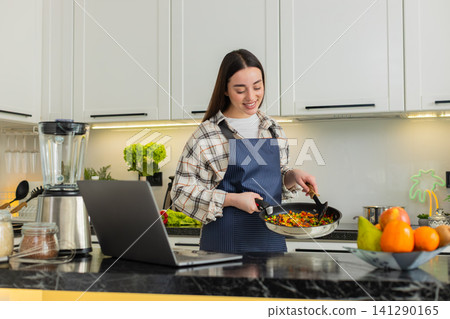 Young woman preparing vegetarian skillet meal checking laptop recipe in cozy home kitchen apartment Young woman preparing vegetarian skillet meal checking laptop recipe in cozy home kitchen apartment 141290165