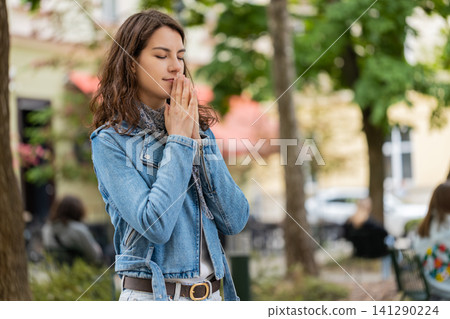 Young woman making prayer gesture walking in city street with peaceful calm sincere emotional mood Young woman making prayer gesture walking in city street with peaceful calm sincere emotional mood 141290224