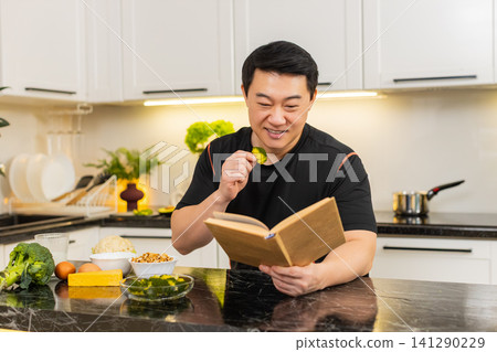 Asian man eats broccoli snack in home kitchen reading book about healthy diet nutrition routine plan Asian man eats broccoli snack in home kitchen reading book about healthy diet nutrition routine plan 141290229